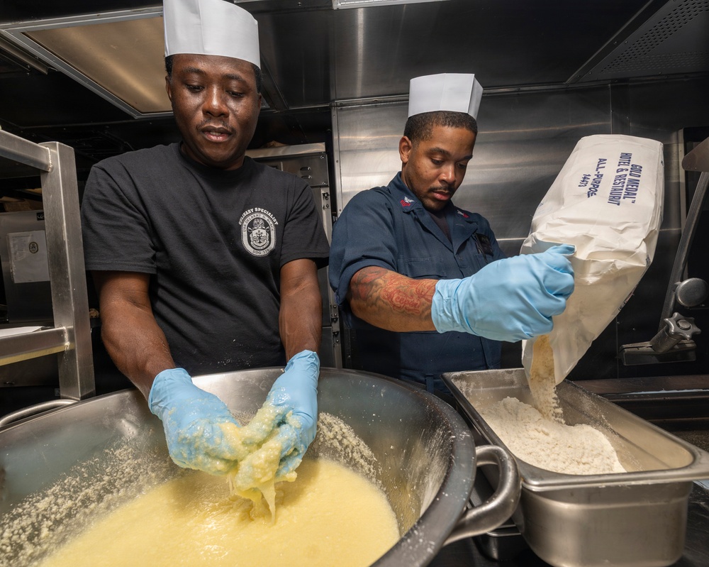 Two military culinary specialists wearing white chef's toques and blue latex gloves are preparing food in what appears to be a naval ship's galley kitchen. The person on the left is mixing a yellow batter or dough by hand in a large metal pan, while the person on the right is pouring flour from a Gold Medal All-Purpose flour bag into a separate metal container. Both are focused on their tasks in a stainless steel commercial kitchen environment.