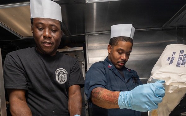 Two military culinary specialists wearing white chef's toques and blue latex gloves are preparing food in what appears to be a naval ship's galley kitchen. The person on the left is mixing a yellow batter or dough by hand in a large metal pan, while the person on the right is pouring flour from a Gold Medal All-Purpose flour bag into a separate metal container. Both are focused on their tasks in a stainless steel commercial kitchen environment.
