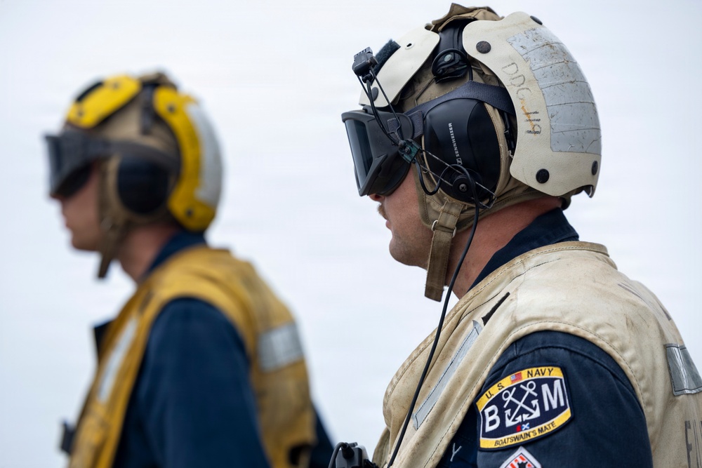 Two U.S. Navy personnel are shown in profile view, wearing flight deck safety gear including helmets and hearing protection headsets. The foreground sailor displays a 'Boatswain's Mate' (BM) rating patch from the U.S. Navy on a tan flight deck jersey. Both individuals appear to be on watch or observing operations, likely on a ship's flight deck.