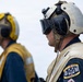 Two U.S. Navy personnel are shown in profile view, wearing flight deck safety gear including helmets and hearing protection headsets. The foreground sailor displays a 'Boatswain's Mate' (BM) rating patch from the U.S. Navy on a tan flight deck jersey. Both individuals appear to be on watch or observing operations, likely on a ship's flight deck.