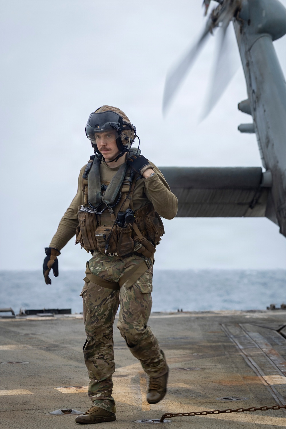 A military personnel member in full tactical gear is walking on what appears to be a naval vessel flight deck with a rotary-wing aircraft (likely an MV-22 Osprey or similar tiltrotor) visible in the upper right. The individual is equipped with a flight helmet, tactical vest, multicam camouflage uniform, and survival/flotation gear appropriate for maritime operations. The overcast sky and open ocean are visible in the background.