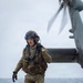 A military personnel member in full tactical gear is walking on what appears to be a naval vessel flight deck with a rotary-wing aircraft (likely an MV-22 Osprey or similar tiltrotor) visible in the upper right. The individual is equipped with a flight helmet, tactical vest, multicam camouflage uniform, and survival/flotation gear appropriate for maritime operations. The overcast sky and open ocean are visible in the background.