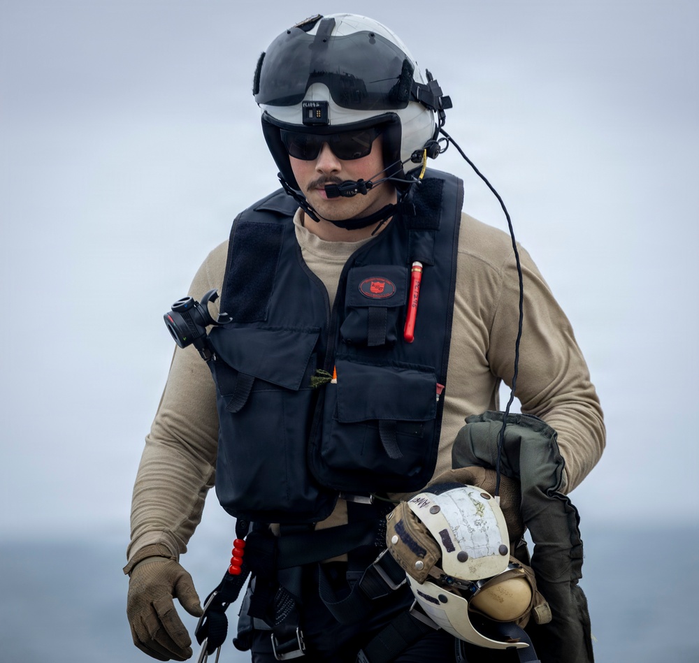 A military aviator or aircrew member is photographed outdoors, likely on a ship deck or flight line, wearing full flight gear. The individual is equipped with a flight helmet, communications headset, survival vest, and tactical gloves, and is carrying a second helmet. The overcast sky and maritime background suggest a naval aviation environment.