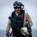 A military aviator or aircrew member is photographed outdoors, likely on a ship deck or flight line, wearing full flight gear. The individual is equipped with a flight helmet, communications headset, survival vest, and tactical gloves, and is carrying a second helmet. The overcast sky and maritime background suggest a naval aviation environment.