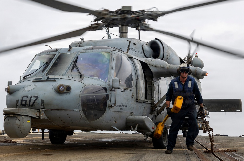 A U.S. Navy helicopter, numbered 617, is shown on what appears to be an aircraft carrier flight deck with its rotors spinning. A naval crew member in blue coveralls, wearing a helmet and eye protection, is walking away from the aircraft carrying yellow chock equipment and chains. Text on the fuselage references 'USS George H.W. Bush' and a commanding officer's name.