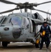 A U.S. Navy helicopter, numbered 617, is shown on what appears to be an aircraft carrier flight deck with its rotors spinning. A naval crew member in blue coveralls, wearing a helmet and eye protection, is walking away from the aircraft carrying yellow chock equipment and chains. Text on the fuselage references 'USS George H.W. Bush' and a commanding officer's name.