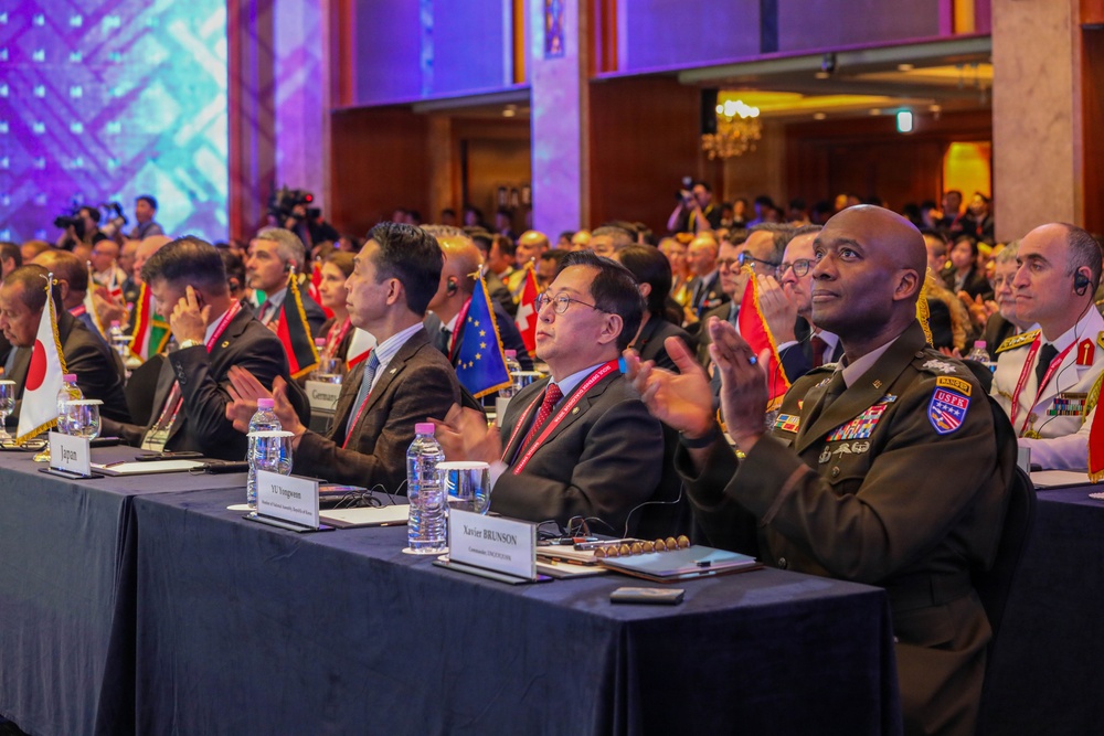 A large multinational military and diplomatic conference is taking place in a formal ballroom setting. Delegates from multiple nations, including a U.S. Army general identified by a nameplate as Xavier Brunson, are seated at a long table adorned with national flags and applauding. The event appears to be a high-level international defense or security forum.