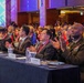 A large multinational military and diplomatic conference is taking place in a formal ballroom setting. Delegates from multiple nations, including a U.S. Army general identified by a nameplate as Xavier Brunson, are seated at a long table adorned with national flags and applauding. The event appears to be a high-level international defense or security forum.