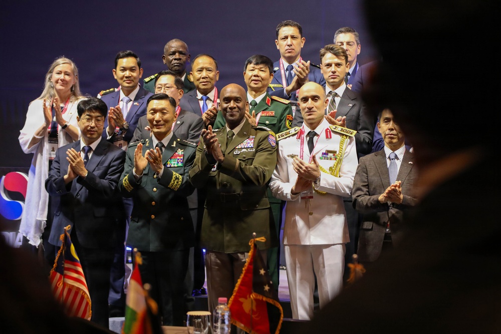A formal multinational military and diplomatic gathering featuring uniformed officers and civilian officials posing together on a stage. Multiple nations appear to be represented based on the variety of military dress uniforms and civilian attire. Several small national flags are visible on a table in the foreground, and a partial South Korean flag emblem is visible on the backdrop.