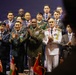 A formal multinational military and diplomatic gathering featuring uniformed officers and civilian officials posing together on a stage. Multiple nations appear to be represented based on the variety of military dress uniforms and civilian attire. Several small national flags are visible on a table in the foreground, and a partial South Korean flag emblem is visible on the backdrop.