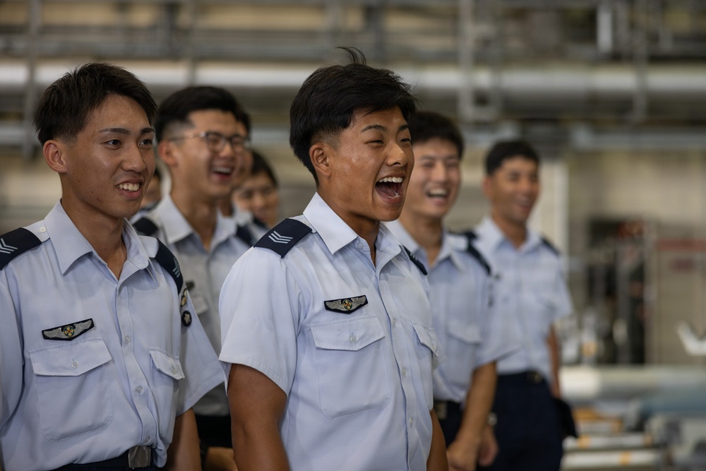 A group of young personnel in light blue military or aviation uniforms are standing in a row, laughing and smiling. The individuals wear wing-shaped badges on their chest pockets and rank insignia on their shoulders, suggesting an aviation or air force context. The mood is visibly joyful and relaxed.