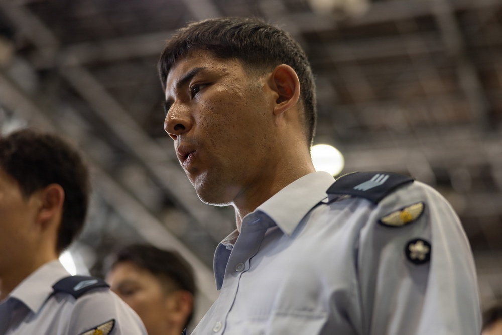 A young man in a light blue military or paramilitary uniform is shown in close-up profile view, appearing to be at attention or participating in a formal event. He has visible perspiration on his face, suggesting physical exertion or heat. Other similarly uniformed individuals are visible in the blurred background.