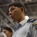 A young man in a light blue military or paramilitary uniform is shown in close-up profile view, appearing to be at attention or participating in a formal event. He has visible perspiration on his face, suggesting physical exertion or heat. Other similarly uniformed individuals are visible in the blurred background.