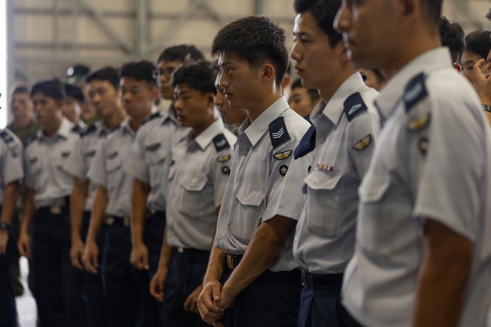 A formation of uniformed military or air force personnel standing in rows inside what appears to be a hangar or large indoor facility. The personnel are wearing light gray short-sleeve shirts with dark navy trousers and display various rank insignia and unit patches on their uniforms. The group appears to be at attention or in a formal assembly posture.