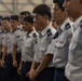 A formation of uniformed military or air force personnel standing in rows inside what appears to be a hangar or large indoor facility. The personnel are wearing light gray short-sleeve shirts with dark navy trousers and display various rank insignia and unit patches on their uniforms. The group appears to be at attention or in a formal assembly posture.