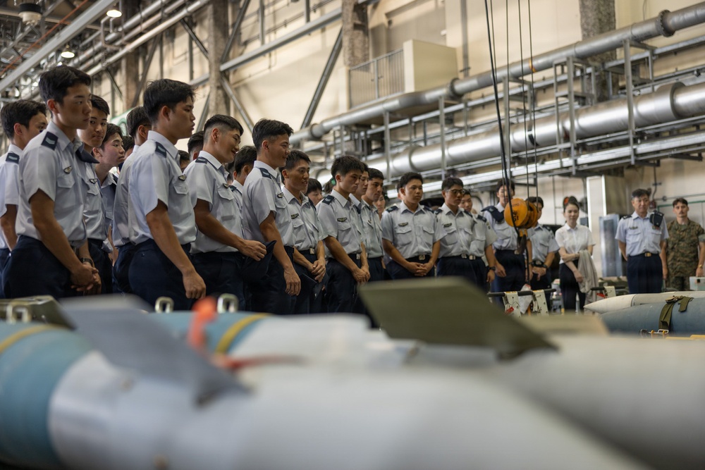 A large group of uniformed military personnel, appearing to be cadets or junior officers in light blue uniforms, are gathered in an industrial hangar or munitions facility. They are standing in a line observing what appear to be aerial munitions or ordnance laid out on the floor in front of them. The setting suggests a training or orientation visit to a weapons storage or maintenance facility.