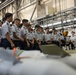 A large group of uniformed military personnel, appearing to be cadets or junior officers in light blue uniforms, are gathered in an industrial hangar or munitions facility. They are standing in a line observing what appear to be aerial munitions or ordnance laid out on the floor in front of them. The setting suggests a training or orientation visit to a weapons storage or maintenance facility.