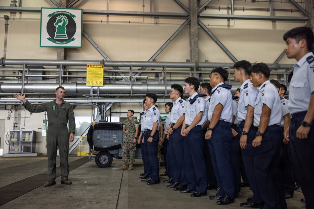 A U.S. Marine Corps aviator in a flight suit is briefing a group of Asian military cadets or officers inside a hangar belonging to VMFA-121 'Green Knights' squadron. The cadets are dressed in white short-sleeve uniform shirts with dark navy trousers and appear to be standing in formation while listening attentively. A second U.S. Marine in camouflage utilities is visible in the background.