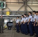A U.S. Marine Corps aviator in a flight suit is briefing a group of Asian military cadets or officers inside a hangar belonging to VMFA-121 'Green Knights' squadron. The cadets are dressed in white short-sleeve uniform shirts with dark navy trousers and appear to be standing in formation while listening attentively. A second U.S. Marine in camouflage utilities is visible in the background.