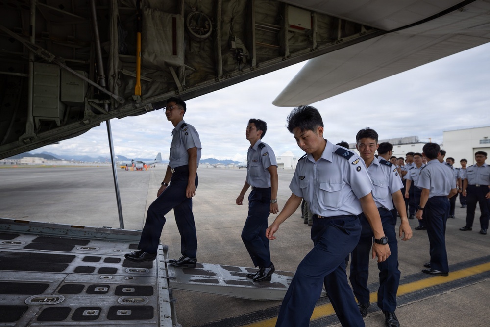 A group of military or air force personnel in light blue shirts and dark navy trousers are boarding or inspecting a large military transport aircraft via its rear cargo ramp. The individuals appear to be of East Asian origin and are wearing matching uniforms with rank insignia visible on some. The setting is an active airfield tarmac with additional aircraft and hangars visible in the background.