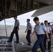 A group of military or air force personnel in light blue shirts and dark navy trousers are boarding or inspecting a large military transport aircraft via its rear cargo ramp. The individuals appear to be of East Asian origin and are wearing matching uniforms with rank insignia visible on some. The setting is an active airfield tarmac with additional aircraft and hangars visible in the background.