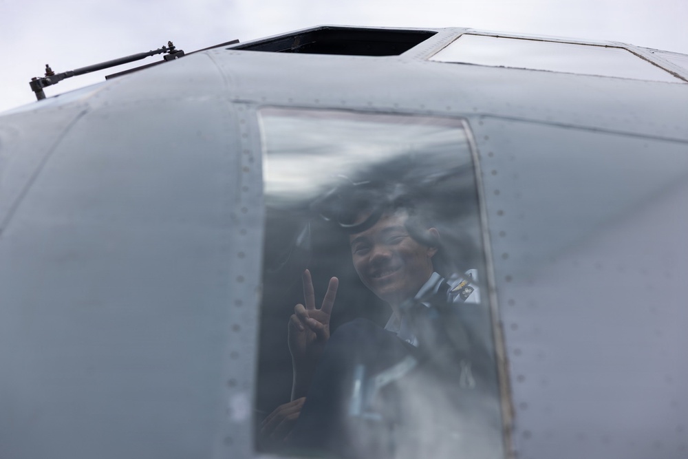 A uniformed military or aviation personnel is visible through the cockpit window of a gray aircraft, smiling and making a peace/victory sign gesture with their hand. The image is taken from a low angle looking up at the aircraft's nose section. The person appears to be in good spirits, possibly during a ground operation or pre/post-flight activity.