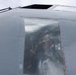 A uniformed military or aviation personnel is visible through the cockpit window of a gray aircraft, smiling and making a peace/victory sign gesture with their hand. The image is taken from a low angle looking up at the aircraft's nose section. The person appears to be in good spirits, possibly during a ground operation or pre/post-flight activity.