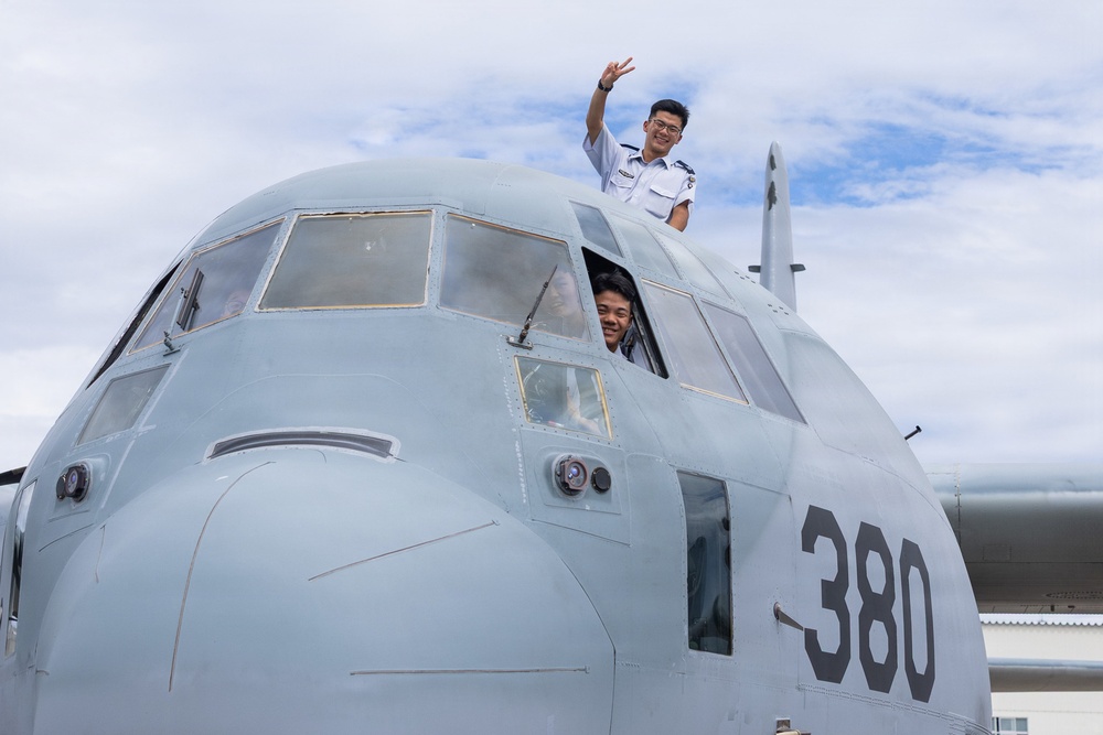 Two individuals in military or aviation uniforms are posing on and around the cockpit of a military transport aircraft marked with the number '380'. One person stands on top of the fuselage making a peace/victory sign, while another leans out of the cockpit window smiling. The aircraft appears to be a C-130 Hercules or similar military transport plane, photographed on the ground under a partly cloudy sky.