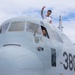 Two individuals in military or aviation uniforms are posing on and around the cockpit of a military transport aircraft marked with the number '380'. One person stands on top of the fuselage making a peace/victory sign, while another leans out of the cockpit window smiling. The aircraft appears to be a C-130 Hercules or similar military transport plane, photographed on the ground under a partly cloudy sky.