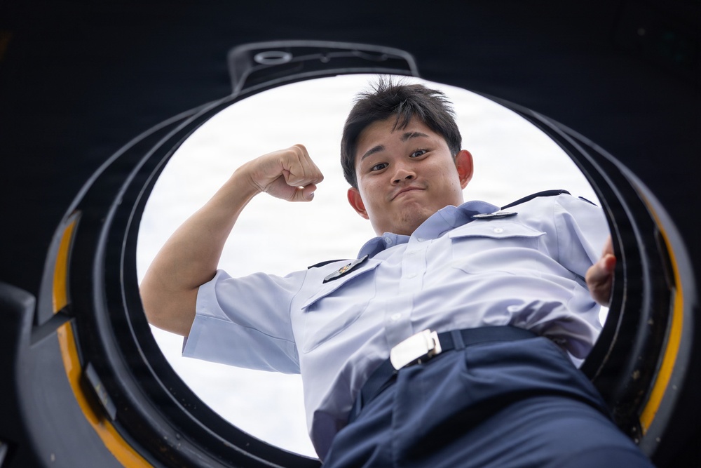 A young person in a light blue military or naval uniform is photographed from below through a circular hatch opening, likely on a vessel or military vehicle. The subject is flexing one arm in a confident pose while looking down into the camera. The low-angle perspective through the circular opening creates a dramatic compositional effect.