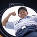 A young person in a light blue military or naval uniform is photographed from below through a circular hatch opening, likely on a vessel or military vehicle. The subject is flexing one arm in a confident pose while looking down into the camera. The low-angle perspective through the circular opening creates a dramatic compositional effect.