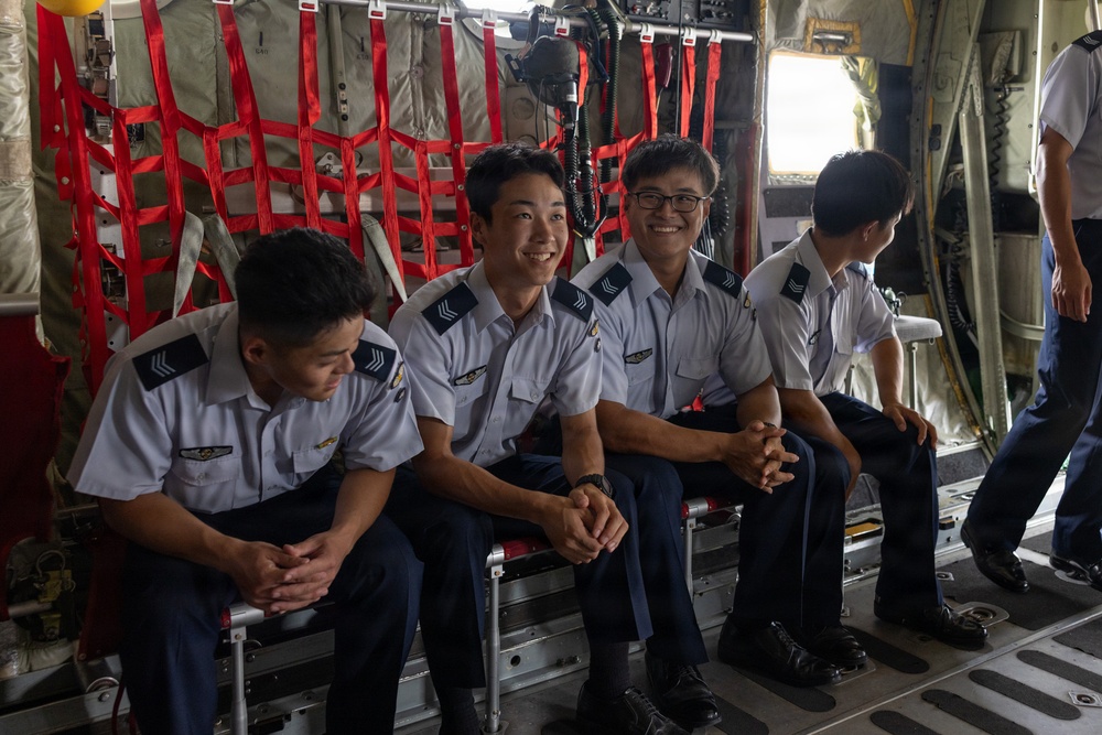 Several uniformed military or air force personnel are seated inside the cargo hold of a military transport aircraft. Three individuals in the foreground are smiling and appear relaxed, while others stand in the background. The uniforms feature sergeant-rank chevrons and wing badges, suggesting an air force context.