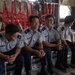 Several uniformed military or air force personnel are seated inside the cargo hold of a military transport aircraft. Three individuals in the foreground are smiling and appear relaxed, while others stand in the background. The uniforms feature sergeant-rank chevrons and wing badges, suggesting an air force context.