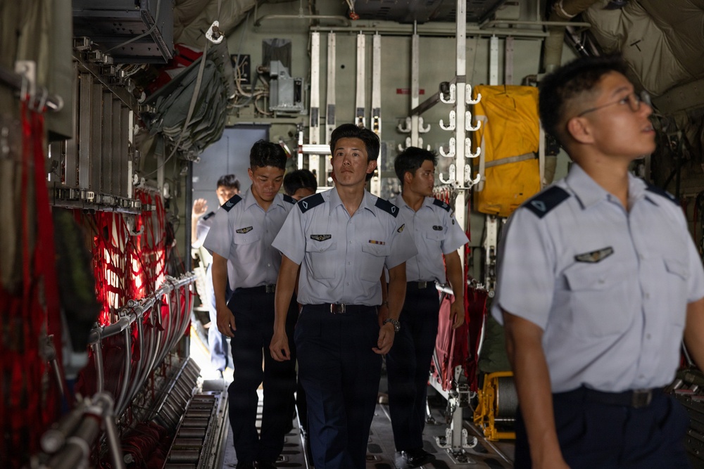 A group of military personnel in light blue uniforms with dark navy trousers are walking through the interior cargo bay of a large military transport aircraft, likely a C-130 Hercules. The individuals appear to be conducting an inspection or tour of the aircraft's interior. Several personnel display wing insignia and rank badges on their uniforms.