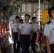 A group of military personnel in light blue uniforms with dark navy trousers are walking through the interior cargo bay of a large military transport aircraft, likely a C-130 Hercules. The individuals appear to be conducting an inspection or tour of the aircraft's interior. Several personnel display wing insignia and rank badges on their uniforms.