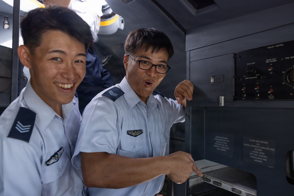 Two individuals wearing light blue military or aviation uniforms with rank insignia and wing badges are examining equipment inside what appears to be an aircraft galley or control area. Both appear to be in good spirits, with one smiling broadly and the other making an expressive face while pointing at a control panel or compartment. The setting suggests a training exercise, tour, or familiarization visit to an aircraft.