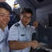 Two individuals wearing light blue military or aviation uniforms with rank insignia and wing badges are examining equipment inside what appears to be an aircraft galley or control area. Both appear to be in good spirits, with one smiling broadly and the other making an expressive face while pointing at a control panel or compartment. The setting suggests a training exercise, tour, or familiarization visit to an aircraft.
