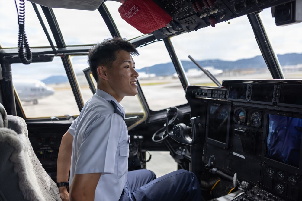 A smiling young male pilot or aviator is seated in the cockpit of a large aircraft, wearing a light blue uniform shirt with a dark tie. The cockpit features numerous instruments, displays, and controls typical of a military or large transport aircraft. The scene is set on an airport tarmac with mountains and other aircraft visible through the cockpit windows.
