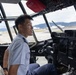 A smiling young male pilot or aviator is seated in the cockpit of a large aircraft, wearing a light blue uniform shirt with a dark tie. The cockpit features numerous instruments, displays, and controls typical of a military or large transport aircraft. The scene is set on an airport tarmac with mountains and other aircraft visible through the cockpit windows.