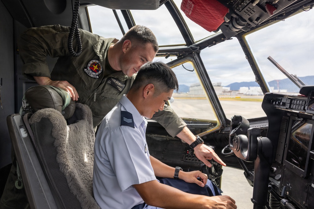 Two military personnel are inside an aircraft cockpit, with one individual in a green flight suit leaning over and pointing at cockpit controls to a second individual seated in a light blue uniform. The interaction appears to be instructional or demonstrative in nature, likely during a tour or training exercise. An airfield with mountains visible in the background can be seen through the cockpit windows.