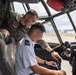Two military personnel are inside an aircraft cockpit, with one individual in a green flight suit leaning over and pointing at cockpit controls to a second individual seated in a light blue uniform. The interaction appears to be instructional or demonstrative in nature, likely during a tour or training exercise. An airfield with mountains visible in the background can be seen through the cockpit windows.