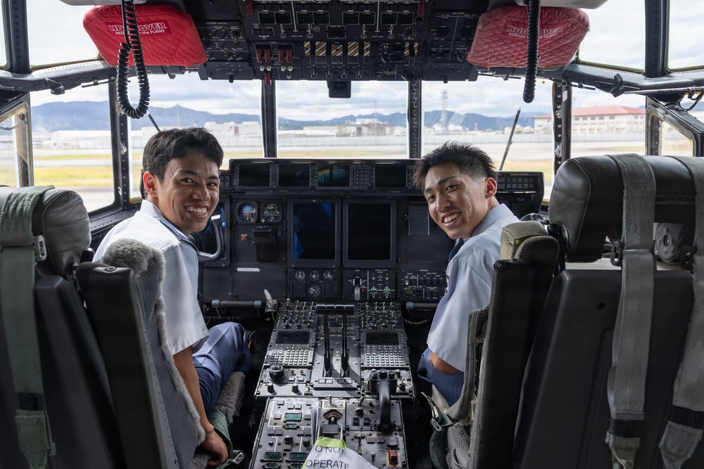 Two young men in white short-sleeve shirts are seated in the cockpit of a large aircraft, smiling at the camera. The cockpit features an extensive array of instruments, displays, switches, and controls typical of a military or large transport aircraft. Red seat covers labeled 'HUD COVER' are visible on the overhead panel, and an airfield with mountains in the background is visible through the windshield.
