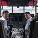 Two young men in white short-sleeve shirts are seated in the cockpit of a large aircraft, smiling at the camera. The cockpit features an extensive array of instruments, displays, switches, and controls typical of a military or large transport aircraft. Red seat covers labeled 'HUD COVER' are visible on the overhead panel, and an airfield with mountains in the background is visible through the windshield.