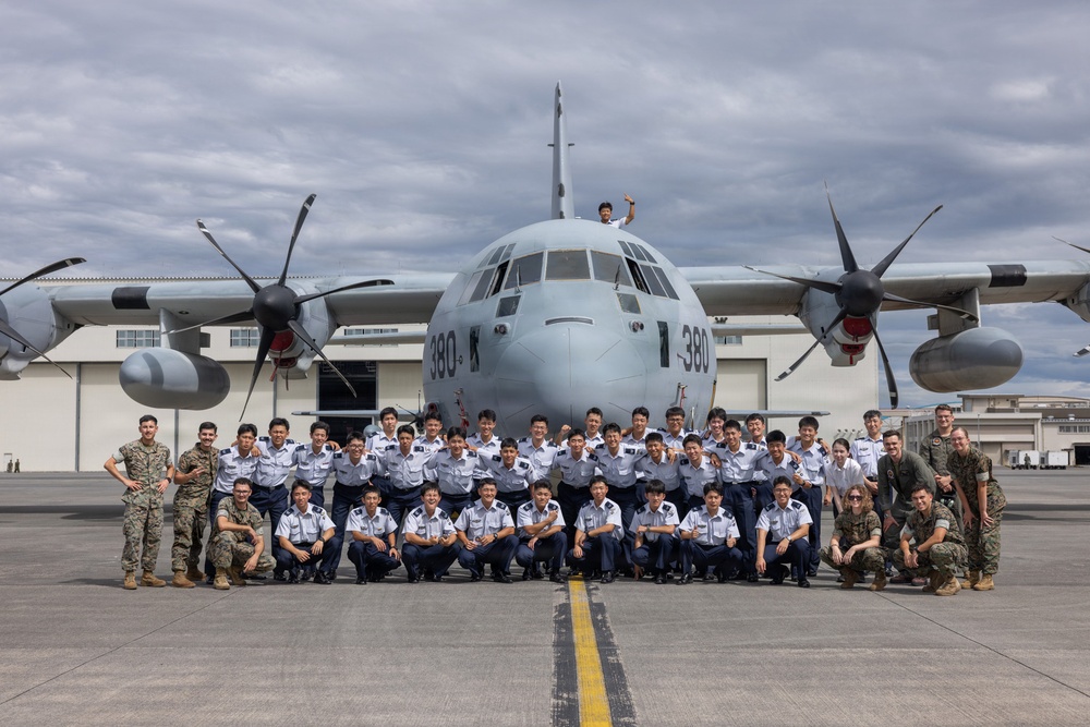 A large group of military personnel pose for a group photograph in front of a four-engine turboprop transport aircraft marked with the number '380' on the tarmac of a military airfield. The group includes personnel in light blue uniforms, likely Japan Air Self-Defense Force (JASDF) cadets or airmen, alongside a smaller number of individuals in U.S. Marine Corps camouflage uniforms. One person is visible standing on top of the aircraft's fuselage near the cockpit.