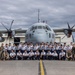 A large group of military personnel pose for a group photograph in front of a four-engine turboprop transport aircraft marked with the number '380' on the tarmac of a military airfield. The group includes personnel in light blue uniforms, likely Japan Air Self-Defense Force (JASDF) cadets or airmen, alongside a smaller number of individuals in U.S. Marine Corps camouflage uniforms. One person is visible standing on top of the aircraft's fuselage near the cockpit.