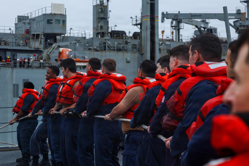 A line of naval personnel wearing navy blue uniforms and bright orange life vests are pulling on a mooring line aboard a military vessel. A large gray naval ship is visible in the background, likely a replenishment or auxiliary vessel. The scene appears to depict a line-handling evolution during a mooring or underway replenishment operation.
