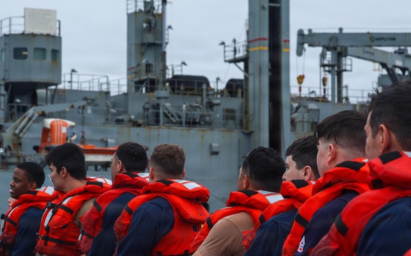 A line of naval personnel wearing navy blue uniforms and bright orange life vests are pulling on a mooring line aboard a military vessel. A large gray naval ship is visible in the background, likely a replenishment or auxiliary vessel. The scene appears to depict a line-handling evolution during a mooring or underway replenishment operation.