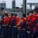 A line of naval personnel wearing navy blue uniforms and bright orange life vests are pulling on a mooring line aboard a military vessel. A large gray naval ship is visible in the background, likely a replenishment or auxiliary vessel. The scene appears to depict a line-handling evolution during a mooring or underway replenishment operation.
