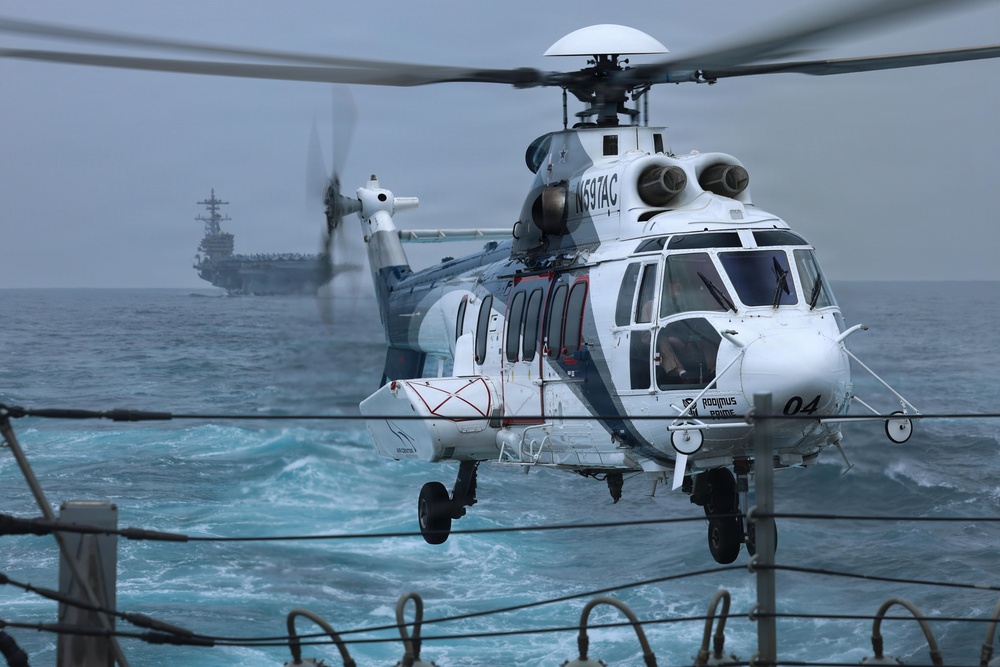 A white civilian helicopter registered N597AC, marked 'Rodimus Prime' with number '04', is hovering or landing on a naval vessel deck over choppy open ocean. In the background, a large aircraft carrier is visible through hazy conditions. The scene appears to depict a helicopter transfer or resupply operation between vessels at sea.