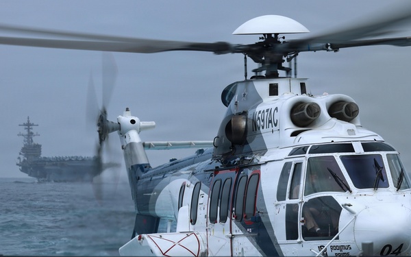 A white civilian helicopter registered N597AC, marked 'Rodimus Prime' with number '04', is hovering or landing on a naval vessel deck over choppy open ocean. In the background, a large aircraft carrier is visible through hazy conditions. The scene appears to depict a helicopter transfer or resupply operation between vessels at sea.